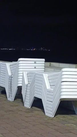 The video shows a nighttime scene along a paved walkway next to a body of water, possibly the sea. Several stacks of white plastic lounge chairs are neatly arranged along the walkway. In the background, across the water, there are distant lights, suggesting a town or city. The sky is dark, and there are some faint lights visible on the horizon. Further down the walkway, a small group of people are visible, walking and talking. The walkway is paved with reddish-brown bricks, and there is a patch of green grass and some palm fronds to the left of the frame. The overall atmosphere is calm and quiet.