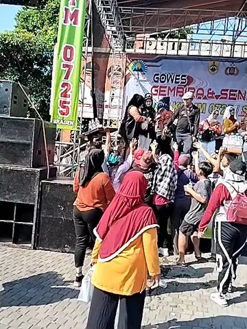 An outdoor scene shows a large crowd of people, including many children and teenagers, gathered around a raised stage. On the stage, several adults are present; one person in a black cap and long-sleeved shirt gestures to the crowd, while another adult in a black shirt appears to be distributing items. Two women in colorful attire are also on stage. Below the stage, the crowd is enthusiastically reaching their hands upwards towards the stage, with some individuals holding up objects. One person in the foreground below the stage holds a tray containing several bottles of water. Behind the stage, a large banner displays text including "GOWES GEMBIRA & SENAM SEHAT" and logos for "EASTLINK" and "BRI". To the left of the stage, a tall green banner reads "0725" vertically and "SRAGEN" at the bottom. Large speakers are visible on the sides of the stage setup. The event appears to be taking place on a sunny day.