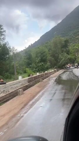 The video captures a view from a moving vehicle traveling along a wet, paved road in a mountainous region. The road surface is extensively covered in standing water and appears muddy in parts, indicating recent heavy rainfall. To the left, a concrete barrier or ditch separates the road from a dense area of trees and lush green vegetation.

In the background, steep, green mountains rise prominently, dominating the landscape under a cloudy sky with hints of blue. Further down the road, a group of people is visible, clustered near several parked cars, including a white truck. Some debris or fallen items, possibly umbrellas, are scattered in the wooded area adjacent to the road. The overall setting suggests a natural, rural environment. The time of day appears to be daytime, with soft, diffused light due to the overcast conditions. No discernible text is visible. The scene is located in Taljarft, Morocco.