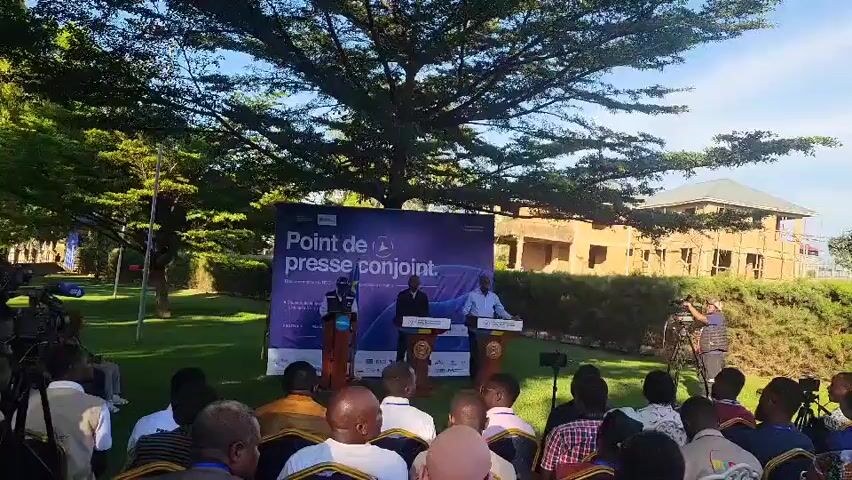 The video shows an outdoor press conference. Three men stand at podiums in front of a backdrop that reads “Point de presse conjoint” which is French for “Joint press conference.” A seated audience faces the speakers. Behind the speakers, a building under construction can be seen, partially obscured by a tree.  A cameraman is also present.