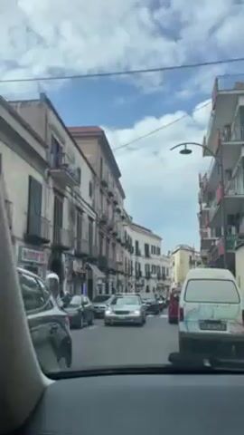 A driver is in a car on a narrow European street. There are cars in front of the driver in the same lane. The buildings on either side of the road have aging façades and balconies. The sky above is partly cloudy.