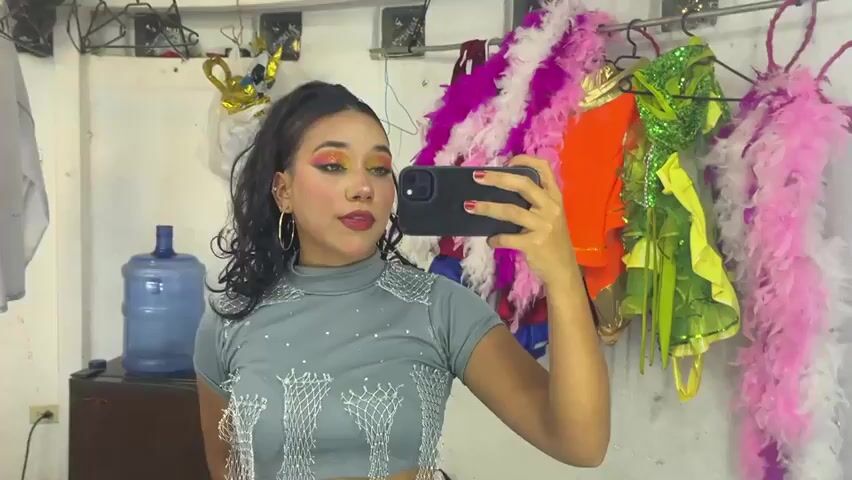 A young woman with dark hair pulled back in a ponytail and heavy eye makeup is taking a selfie in a room that appears to be a backstage area or dressing room. She is wearing a grey, high-necked top with reflective embellishments. She holds a black smartphone in her right hand, aiming it at herself. Behind her, on the wall, are various colorful costumes and accessories, including pink and white feather boas, and green and yellow sequined outfits hanging on hooks. To the left, there is a large blue water jug and a dark cabinet. The overall impression is one of preparation for a performance or event.