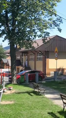 The video shows a red car parked next to a building. A person is riding a bicycle towards the car. The building appears to be a school or a community center. There are trees and benches in the foreground. The weather is sunny and clear.