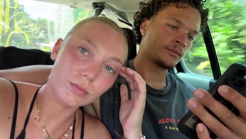 A young couple is in a car. The woman, blonde with blue eyes, is in the foreground, holding a phone up to record a selfie video. She is wearing a black tank top and a gold necklace. The man, with curly brown hair and a mustache, is next to her, smiling and holding a phone. They are driving through a lush, green area. The woman makes a kissing face. The camera is shaky.