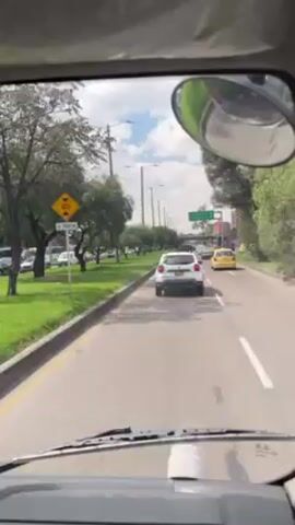 The video shows a street in Bogota, Colombia, from the perspective of a driver in a large white vehicle. The camera is inside the vehicle and faces forward through the windshield. A white car and a yellow taxi are driving on the street, and there are trees and buildings on either side. The view in the side mirror shows the driver, a young man with glasses, a tattoo on his arm, and a seatbelt, taking a photo with his phone. The weather appears to be sunny.