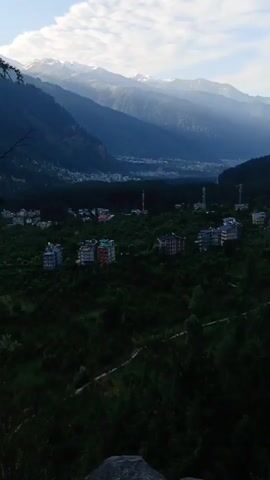 A panoramic view of a mountainous landscape. In the foreground, dense green foliage covers the hills, dotted with several multi-story buildings with red, white, and blue facades. Several communication towers are visible amongst the trees. In the mid-ground, a town or city is nestled in a valley, with more buildings and infrastructure visible. The background features a series of majestic, snow-capped mountains under a bright, partly cloudy sky. Sunlight casts rays across the mountain peaks, highlighting their rugged contours. The overall scene suggests a serene and expansive natural environment with human settlements integrated into the landscape.