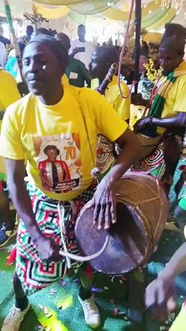 Here's a detailed description of the video:

The video captures a vibrant scene of a group of men performing traditional music, likely at a celebration or event. The main focus is on a man playing a large drum. He's wearing a yellow t-shirt with a graphic print, a black head covering, and a colorful patterned skirt. He is positioned in the center of the frame, striking the drum with his hands to create rhythm.

Surrounding him are other men, also dressed in yellow t-shirts. Some of them are holding long sticks. The background shows a venue decorated with light green and yellow fabrics. Confetti is scattered across the floor, adding to the celebratory atmosphere.