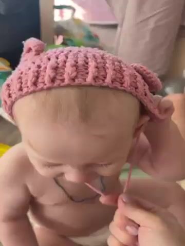 The video shows a baby taking off a pink knit hat with bear ears. The baby is bare-chested and wears a blue necklace with a pendant. The baby removes the hat string from under their chin, removes the hat completely, and turns to face the camera, touching their face with their hand. There are toys and furniture visible in the background. A person's hand is visible as they remove the hat string.