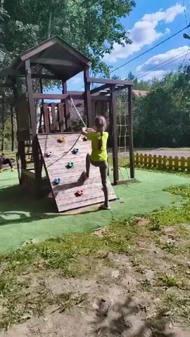 The video shows a young girl in a yellow t-shirt and grey pants climbing up a wooden climbing wall on a playground. She is using a rope to assist her ascent. The climbing wall is attached to a larger wooden play structure that includes a small tower with a roof and a net for climbing. The playground has a green artificial turf surface with some patches of dirt and grass visible. Trees and a fence can be seen in the background. The sky is blue with some clouds. Another person, partially visible and appearing to be a child, is sitting on the ground to the left of the frame. The lighting suggests it is daytime.