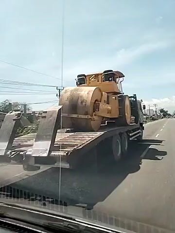 The video shows a yellow Sakai brand road roller being transported on a flatbed truck. The truck is traveling on a road with utility poles and buildings in the background. The view is from inside a car, looking out the windshield.
