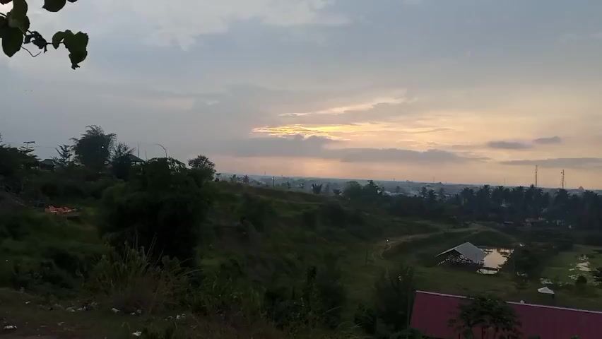 The video presents a wide-angle view of a landscape at dawn or dusk. The sky is partially obscured by clouds, with the sun appearing to peek through a gap. The foreground is dominated by a grassy hill with various trees and vegetation. In the middle ground, there's a small structure with a white roof on the edge of a body of water.  Behind this, a flat plain stretches into the distance, featuring dense foliage and scattered buildings, with some tall antenna-like structures. The overall atmosphere is calm and serene, suggesting a peaceful natural setting at a transition point in the day.