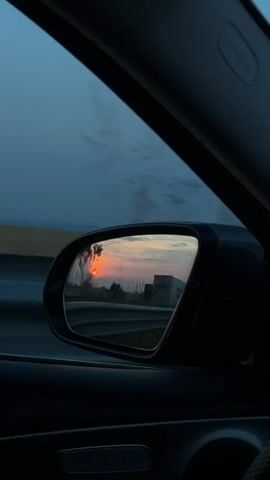 The video captures a first-person view from inside a moving vehicle, looking towards the passenger side. The dark interior of the car, including a portion of the door panel and an "AIRBAG" label above the window, is visible in the foreground. The car's passenger side mirror reflects a sunset, showing a distinct orange sun low on the horizon, partially obscured by the dark silhouettes of trees. A road barrier is also visible in the mirror's reflection. Through the car window, the external environment appears blurred, indicating motion, with a dark blue-grey sky.