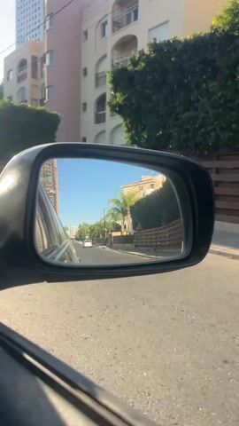 The video captures a view from a car, primarily focusing on the side-view mirror. The mirror reflects a street scene with buildings and trees. In the background, a tall, modern building with glass windows is visible, along with other residential buildings featuring balconies and arches. Green trees and hedges line the street, creating a somewhat lush urban environment. The road surface appears to be asphalt. Several vehicles are seen in the reflection of the mirror, including what looks like a white pickup truck and a white SUV. Sunlight is casting shadows, indicating it is likely daytime. The perspective suggests the car is moving forward, and the video shows a short duration of this movement.