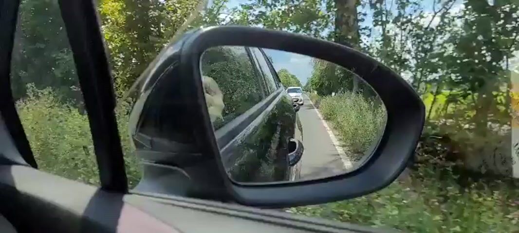 A point-of-view shot from inside a moving car shows a side-view mirror reflecting a sunny rural road. The mirror shows the rear passenger-side of the car, which is black. Reflected in the mirror, the road ahead is visible, with a white car in the distance. The road is lined with green foliage. The car is moving, as indicated by the passing scenery.