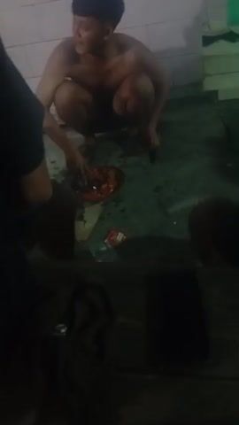 A young male, appearing to be of East Asian descent, is crouched down on a damp, possibly tiled, floor. He is shirtless and has dark hair styled in a buzz cut. He is interacting with a shallow, round dish filled with a reddish-orange food item, possibly a chili paste or sauce, using his hands to mix it. To his left, a portion of another person's arm and shoulder is visible, indicating the presence of at least one other individual. On the floor near the dish are several small packets, one of which appears to be a food packet with red and white packaging. The environment seems to be indoors, with a tiled wall in the background and a green grate-like object visible to the right. The lighting is dim, casting shadows on the scene. The overall impression is of a casual, possibly communal, meal preparation or consumption in a somewhat utilitarian setting.