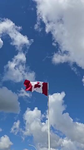 The video shows a Canadian flag waving on a flagpole against a bright blue sky with scattered white clouds. The camera angle is looking up at the flag. The flag is predominantly red and white with a maple leaf emblem in the center. The pole is a light color, likely metal. The lower part of the frame shows a hint of a light-colored curved object, possibly a street lamp or fence. The overall impression is a clear, sunny day.