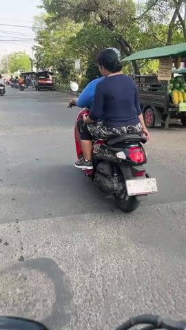 A first-person perspective shot captures two individuals riding a scooter down a paved road. The rider is a person wearing a blue shirt and dark pants, and the passenger, positioned behind the rider, is wearing a dark shirt with a patterned waistband and dark pants. The scooter is dark colored and features a visible license plate. The road appears to be in an urban or semi-urban setting, with other vehicles, including scooters and what appear to be tuk-tuks, visible in the background. Trees and foliage line the side of the road, and the sky is visible above, suggesting an outdoor daytime scene. The perspective suggests the camera is mounted on the handlebars of another scooter.