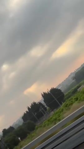 The video shows a view from a moving vehicle, possibly a train or car, looking out at a landscape under a cloudy sky. The sky is a mix of grey clouds with some shafts of sunlight breaking through, creating a dramatic effect. The landscape consists of trees and greenery, with some power lines and a utility pole visible on the left side of the frame. The camera is tilted at an angle, giving a dynamic perspective. The overall impression is of a journey through a somewhat overcast but picturesque environment.