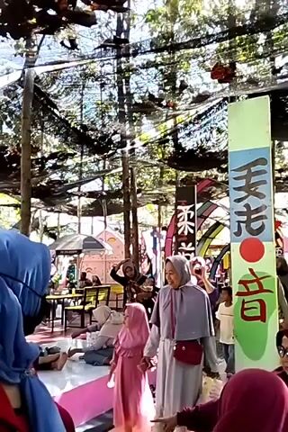 The video shows an outdoor area with several people, including children and adults, enjoying a day out. The environment appears to be a playground or recreational area, with a covered structure providing shade. Various colorful decorations and structures are visible, including a tall, green pillar with Japanese writing and a red circle, and a large, white, heart-shaped object with colorful designs. Hammocks are seen hanging between trees, and a slide is present in the background. People are seen walking, interacting, and sitting. Some individuals are wearing hijabs, suggesting a cultural context. The overall atmosphere appears to be relaxed and family-friendly.
