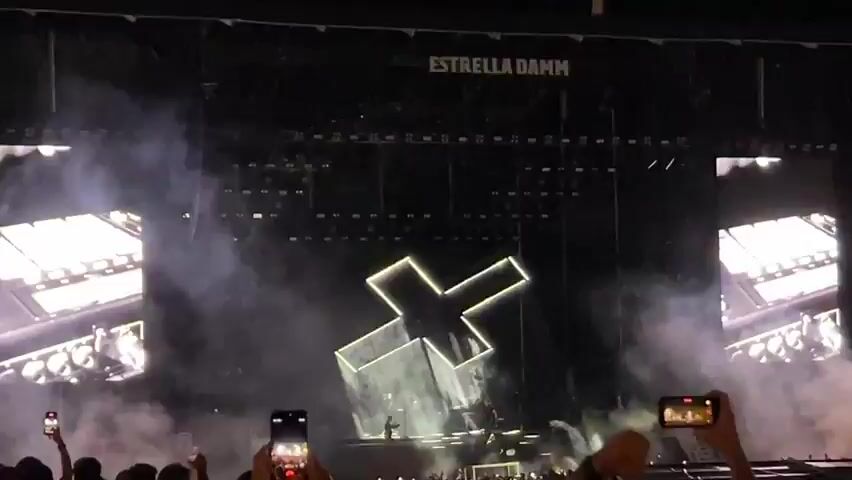 The video shows a live concert. The stage has a large, illuminated "X" shape in the center, with smoke effects and bright stage lights. On either side of the stage are large screens displaying close-ups of a man's face. The audience is visible in the foreground, with many people holding up their smartphones to record the performance. The word "ESTRELLA DAMM" is displayed prominently on a banner above the stage.