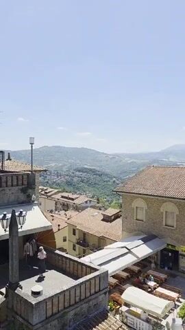 The video shows a panoramic view of a hilly landscape in Italy, presumably in or near San Marino. The camera slowly pans right, revealing buildings with tile roofs, including a bar with outdoor seating. In the background, a vast expanse of rolling hills stretches towards the horizon under a clear blue sky. The foreground features a low stone wall and trees, further framing the scenic view.
