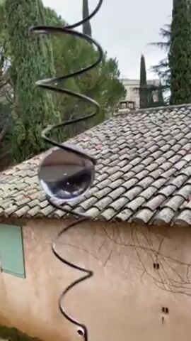 The video shows a metal spiral with a glass ball in it. The ball is moving slowly down the spiral, seemingly powered by rain falling from above. In the background is a weathered building with a tiled roof and a green shuttered window, framed by green trees under a grey sky.