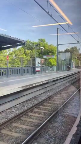 The video shows a train departing from or passing through an outdoor station platform on a sunny day. The view is from inside the moving train, looking out a window towards the platform and the tracks.

On the concrete platform, a person is briefly visible near a small, modern glass structure, possibly an elevator or waiting shelter, which has "2A" written on it. Several lampposts are spaced along the platform, and various advertising boards are displayed. A red circular sign with a white "T" and an arrow, characteristic of a transport system logo, is prominently visible. Further down the platform, a red circular sign with a white "X" (prohibition symbol) is seen.

The platform is bordered by a metal fence, behind which lush green trees and foliage are abundant, suggesting a semi-rural or suburban setting. Overhead catenary lines and power poles span the tracks, indicating an electric train system. The tracks themselves are laid on ballast, with a second set of tracks visible parallel to the platform. The sky is bright blue with scattered clouds. The train moves steadily, showing more of the fenced-off natural area as it progresses.