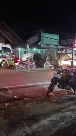 The video shows a nighttime street scene. A red dump truck with a yellow cab is driving past a row of buildings. There are several motorcycles parked on the side of the road, and people are gathered outside one of the buildings. The street is paved, and there is debris on the side of the road. The lighting is dim, with the primary illumination coming from the headlights of the vehicles and the lights from the buildings. A white truck with colorful graphics is parked on the left side of the frame.