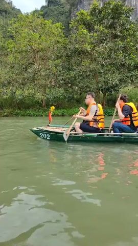 The video shows a first-person perspective of someone paddling a boat on a river. In front of the paddler, another long, narrow boat is being rowed by two men wearing yellow life vests. A woman in a traditional conical hat is also in the boat, guiding it with an oar. The river is bordered by lush green trees and tall rock formations can be seen in the background.  The boat in front has the number "202" painted on its side and is decorated with a small orange flag.