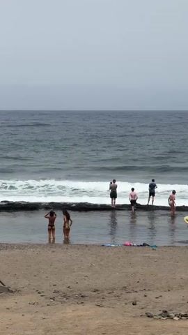 The media file depicts a serene beach scene in Arona, Spain, under an overcast sky, suggesting it's likely morning or late afternoon. The calm, grey ocean stretches to the horizon, with gentle waves lapping against a dark, rocky shoreline.

Several individuals are present, enjoying the coastal environment. On the sandy beach in the foreground, two young women in bikinis stand near the water's edge. To their right, a person dressed in black carries a yellow surfboard, walking away from the water.

Further back, a line of people stands on the rocky formation separating the shallow pool from the open sea. One person is holding up a phone, seemingly taking a photo. Others are observing the water or interacting with each other. The overall atmosphere appears relaxed and tranquil, with people enjoying a quiet day at the beach.

There are no visible text elements within the media file. The location context is Arona, Spain.