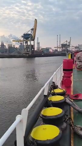 The video begins from the perspective of someone on a ship, looking out towards an industrial waterfront. In the foreground, the railing of the ship is visible, along with several large, yellow-capped bollards. The water is calm with slight ripples. In the background, a large industrial complex is visible. Several tall cranes, some yellow and blue, dominate the skyline, along with numerous smokestacks emitting plumes of smoke. Various industrial buildings and structures are scattered across the waterfront. A few barges or smaller boats are also present on the water near the industrial complex. The sky is overcast with some visible clouds. The overall scene depicts a busy industrial port area, likely during sunrise or sunset due to the lighting conditions.