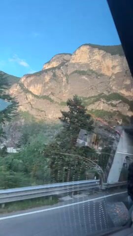 The video shows a view from a vehicle, possibly a train or a bus, moving through a mountainous landscape. The camera is positioned to capture the scenery through the windows. In the foreground, the metallic structure of the vehicle's interior is visible, including windows and some lighting fixtures. Outside, a bridge structure with distinctive white supports is prominent. Beyond the bridge, rugged, rocky mountains rise against a clear blue sky. A tall green tree stands to the left of the frame. The vehicle appears to be in motion, as indicated by the passing scenery and the reflections on the windows. The overall impression is one of travel through a scenic, natural environment.