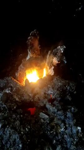 In a dimly lit, almost black environment, a small fire burns brightly. The flames are a vivid orange and yellow, contrasting sharply with the surrounding darkness. The fire is contained within a structure of rough, dark gray rocks or debris, which seem to have a porous, almost sponge-like texture. The edges of these materials appear jagged and irregular. The ground around the fire is similarly dark and rocky. The perspective is a close-up, focusing on the fire and its immediate surroundings, with the background receding into an undifferentiated darkness.