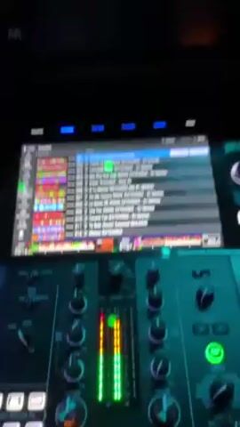 Here's a description of the video:

The video shows a DJ booth overlooking a crowded dance floor. The DJ console is in the foreground, featuring a digital display and control knobs, with lights on the console. A screen displays a song list and audio waveforms. The camera pans up to reveal a nightclub scene with a large crowd of people dancing under colorful lights. The nightclub, which appears to be called "The Tango," is filled with people in casual attire, illuminated by neon lights overhead. The environment is energetic and upbeat.