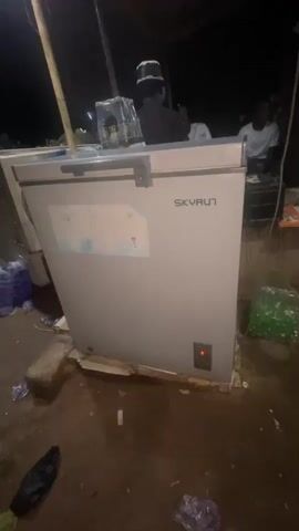 A chest freezer is visible in the foreground, with the brand name SKYRUN on its lid. There is a red indicator light on the front of the freezer. The freezer is placed on pieces of wood on a dirt surface. Several plastic bottles and bags are scattered around the freezer. In the background, a few people are partially visible. One person wearing a white shirt is seated behind what appears to be a counter or table. Another person is standing behind the freezer, facing away from the camera. The scene appears to be outdoors, possibly at a market or roadside stall, given the dirt ground and scattered items. The lighting suggests it is nighttime or dusk.