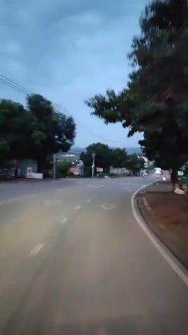 This video captures a point-of-view perspective, likely from a vehicle, traveling down a paved road in Pờ Y, Vietnam. The scene is a rural or suburban road lined with trees on both sides, with buildings and houses visible in the distance. Power lines crisscross the sky. The road is marked with white and yellow lines, including a crosswalk with white stripes and a diamond-shaped marking. Several vehicles are on the road, including cars and possibly motorcycles. A few people are visible on the sidewalks and near the buildings.

The time of day appears to be dusk or early evening, as indicated by the dim, cloudy sky and the headlights of the vehicles. The weather seems to be overcast. The overall mood is peaceful and transitional, suggesting the end of the day. There is no discernible text in the video. The location cues, such as the style of buildings and road markings, are consistent with a Vietnamese setting.