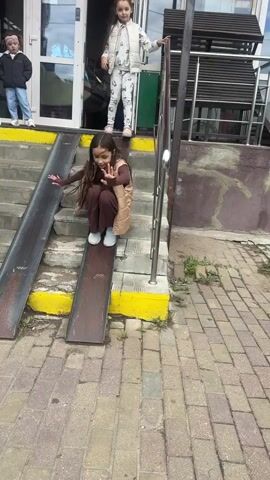 A group of young girls are playing on a ramp that is attached to a set of stairs. The ramp is made of metal and has yellow markings on the sides. The stairs are made of concrete and are also marked with yellow. The girls are all wearing casual clothing. One girl is sliding down the ramp, while another is standing at the top of the ramp and watching. Another girl is standing on the stairs and appears to be waiting for her turn. The environment appears to be outdoors, with a brick pathway in front of the stairs and a building in the background. There are also some plants growing near the building.