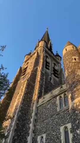 The video shows a church building from a low angle. The church is constructed of gray stone. The building has a tall, pointed tower, a shorter, round tower, and a large arched entrance with windows above. The sky is a clear blue. The sun is shining and casting a golden light on the stone.