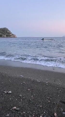 The video shows a beach with dark sand and pebbles in the foreground. Gentle waves with white foam are rolling onto the shore. In the background, the blue sea stretches out towards a rocky island on the left and a hazy horizon on the right. A small white boat with an outboard motor is anchored in the water, slightly off-center to the right. The sky is a soft, pale gradient of purple and pink, suggesting either sunrise or sunset. The overall scene is tranquil and natural.