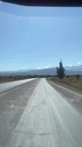 The video captures a view from inside a vehicle traveling on a paved road. The road is partially wet and appears to have some wear and tear, with visible cracks. To the left, there is another lane of the road, also appearing wet. A dark-colored car is seen in the left lane, moving away from the camera. To the right of the road, there is dry, grassy terrain with a lone, tall, slender tree standing upright. In the distance, a mountain range stretches across the horizon under a clear, bright blue sky. Power lines are visible running across the sky. The overall environment suggests an outdoor setting on a sunny day, possibly in a rural or semi-arid region.