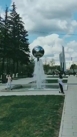 The video shows a park setting on a sunny day with scattered clouds. The camera is positioned on a paved walkway, looking towards a fountain with a large, reflective sphere on top. Water is spraying from the fountain. To the right of the fountain is a tall, abstract monument. Several people are visible in the park, including two children, one on a scooter and the other walking along the edge of the fountain. There are trees lining the perimeter of the park. The setting appears to be a public park in an urban area.