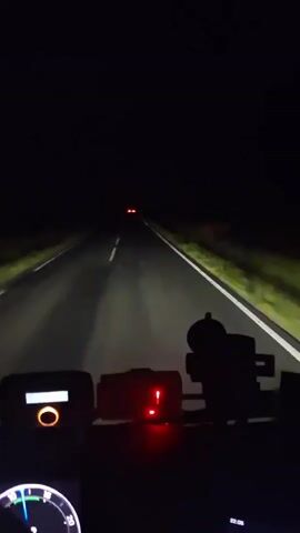 The video shows a first-person perspective from inside a vehicle driving at night on a dark, rural road in Corlățel, Romania.

In the initial frames, the vehicle's headlights illuminate the road ahead, showing white solid and broken lines. On the dashboard, two devices are visible; one has an orange circular light, and another displays two red indicator lights. The road stretches into the darkness, with distant red lights of another vehicle barely visible.

At the 0:02 mark, a sudden, bright flash of lightning illuminates the entire sky and the surrounding landscape. The sky turns a pale purple-white, revealing vast open fields on either side of the road and several power lines in the distance. Raindrops are visible on the windshield, indicating a thunderstorm. The road surface appears wet under the lightning's glow.

The lightning flash quickly subsides, returning the scene to deep night by 0:03. The vehicle continues driving, with only its headlights piercing the darkness and the red and orange lights on the dashboard providing illumination inside the cabin. The distant red taillights of the preceding vehicle remain visible. No specific text is clearly legible on any of the dashboard devices.