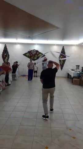 In an indoor hall, likely a community center or event space, a group of people are gathered. The room has tiled floors and white walls, with decorative wall hangings featuring floral arrangements. A suspended ceiling with recessed lighting illuminates the scene.

The central activity involves a man in a black t-shirt and light-colored pants performing with a long, thin, purple ribbon. He is twirling it in wide arcs around himself. In the background, a woman in a floral blouse and blue pants stands near a young boy in a white shirt and shorts. Another man, wearing a grey shirt and pants, stands behind the woman and boy, holding a red object. Several other individuals are seated on chairs along the left side of the room, observing the performance. Tables are set up along the right wall, one covered with a white tablecloth, suggesting a meal or refreshment area.

The lighting appears to be artificial, indicating the event is likely taking place in the evening or during the daytime indoors. The atmosphere seems festive, with a sense of entertainment and social gathering. The location cues, such as the decor and the overall setup, are consistent with a rural community event in Belarus. There is no visible text in the media file.