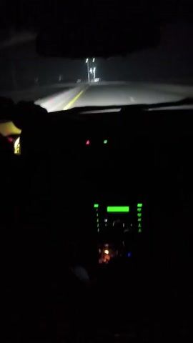 The video shows a view from inside a car driving on a highway at night. The car is moving forward, illuminated by its headlights.  Streetlights are visible along the side of the road. Inside the car, the dashboard is partially visible, with green lights from the radio and other controls glowing. The driver's hands can be seen on the steering wheel. A rear-view mirror is positioned in the upper middle of the frame. A voice is heard shouting, accompanied by engine noise.