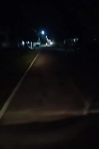 The video shows a road at night, illuminated by streetlights and the headlights of a vehicle. The road appears to be paved and has a white line on the left side. On the right side of the road, there are some buildings or structures, and on the left side, there is vegetation. There is a vehicle with its headlights on, approaching from the distance. The overall scene is dark, with the lights creating pools of illumination on the road and surrounding areas. The camera appears to be mounted on a moving vehicle, likely in the front.