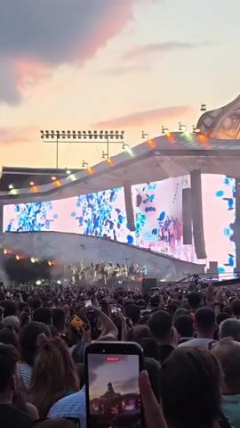 The video shows a wide shot of a large outdoor concert at dusk. A massive stage with multiple screens is visible, displaying bright, abstract graphics with the text "FLORIDA" prominently featured. Several figures are on stage, appearing to be performers. In front of the stage, a vast crowd of people are gathered, many holding up their smartphones to record the event. The sky above is a warm gradient of orange and pink, indicative of sunset. The overall atmosphere is that of a lively musical performance with a large audience.