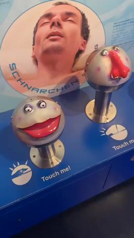 The video shows an interactive exhibit with two stylized face sculptures on a blue counter. The sculpture on the left has a large, red, open mouth with white teeth and large, round, blue eyes. The sculpture on the right has a large, red, bulbous nose and smaller, round, red eyes. Above the sculptures is a large circular image of a man's head with his eyes closed, and text that reads "SCHNARCHEN" and "SNORING". The text "Touch me!" is visible below each sculpture. In the second clip, a hand with pink painted fingernails reaches out and touches the sculpture on the left. The video pans to the right, revealing a third sculpture with a red nose and eyes, and then a fourth sculpture with a smiling mouth and eyes. The text "NIESEN" and "SNEEZING" is visible on the wall behind the sculptures, along with an image of a person sneezing. There are also images of bubbles on the wall. The environment appears to be a museum or science center.