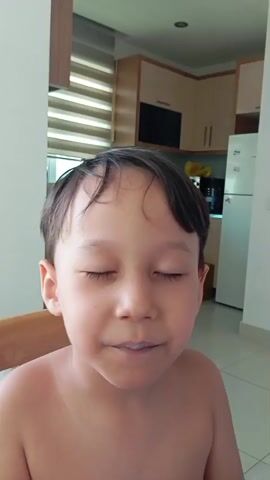 The video shows a close-up of a young boy's face. He is shirtless and has dark hair. His eyes are closed, and he is sticking out his tongue, which is colored a vibrant blue. The background appears to be a kitchen or dining area, with cabinets and a refrigerator visible. The lighting suggests it is daytime. The boy's expression is playful. There is no visible text in the video.