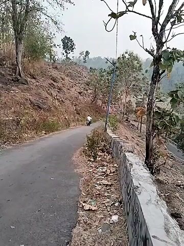A motorcycle is seen driving down a paved road that winds through a hilly, sparsely vegetated landscape. The road is flanked by a low stone wall on the right side, and the terrain slopes upwards to the left, covered in rocks and dry brush. The motorcycle is white and appears to be carrying a single rider. The background features more trees and distant hills, with a slightly overcast sky. There is some debris visible along the roadside, including plastic bags and dried leaves. The scene suggests a rural or undeveloped area.
