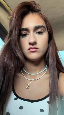 A young woman with long, brown hair is shown from the chest up in a close-up shot. She is wearing a white tank top with black polka dots and two necklaces. One necklace is made of small blue and white beads, and the other has a small gold pendant. She has on makeup, including eyeshadow and lipstick. Her hair is partially obscuring her face. She makes a few facial expressions, including puckering her lips and looking at the camera. The background is blurred but appears to be indoors with a light blue wall visible.
